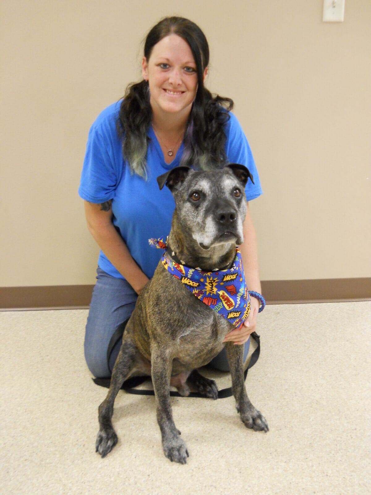 Anlee kneeling beside a senior brindle dog in a comic-print bandana